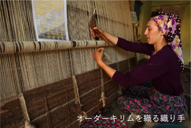 縦糸を張った垂直機（ハタ）の前でキリムを織る女性の織り手の写真